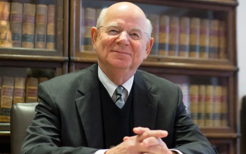 Elderly man in a suit sits at a desk with law books behind him, smiling slightly.