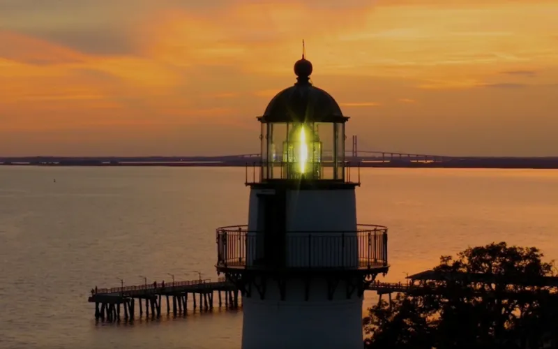 Lighthouse at sunset with calm water and vibrant orange sky.