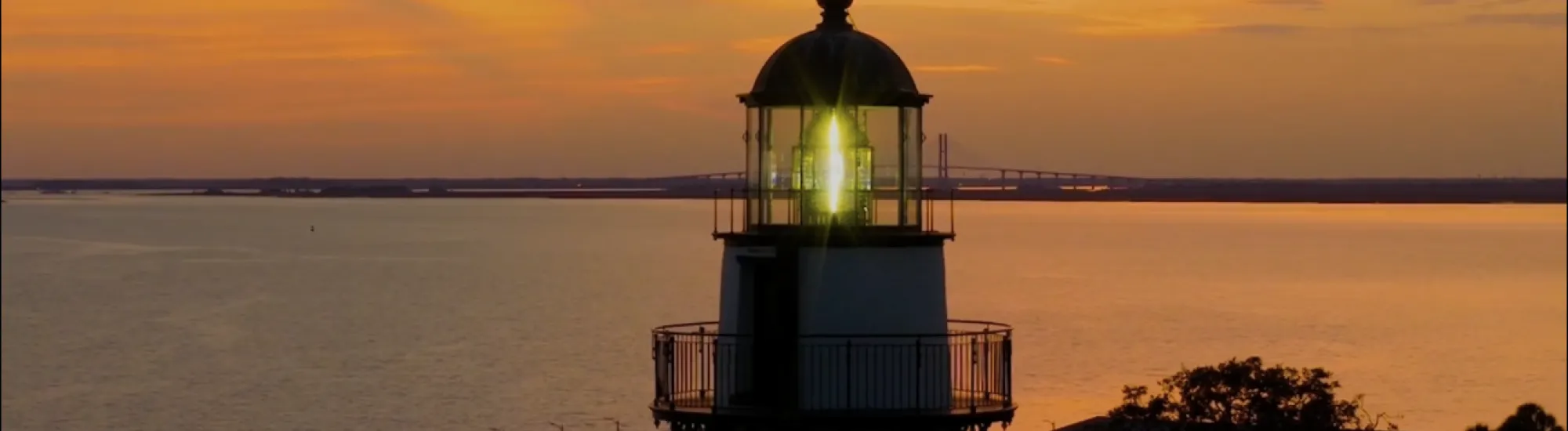Lighthouse at sunset with calm water and vibrant orange sky.