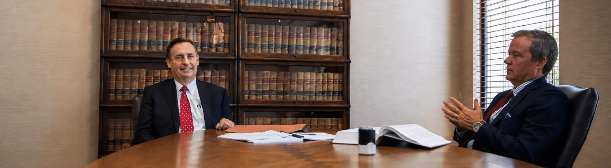 Two men in suits talking at a large wooden table in an office with law books.